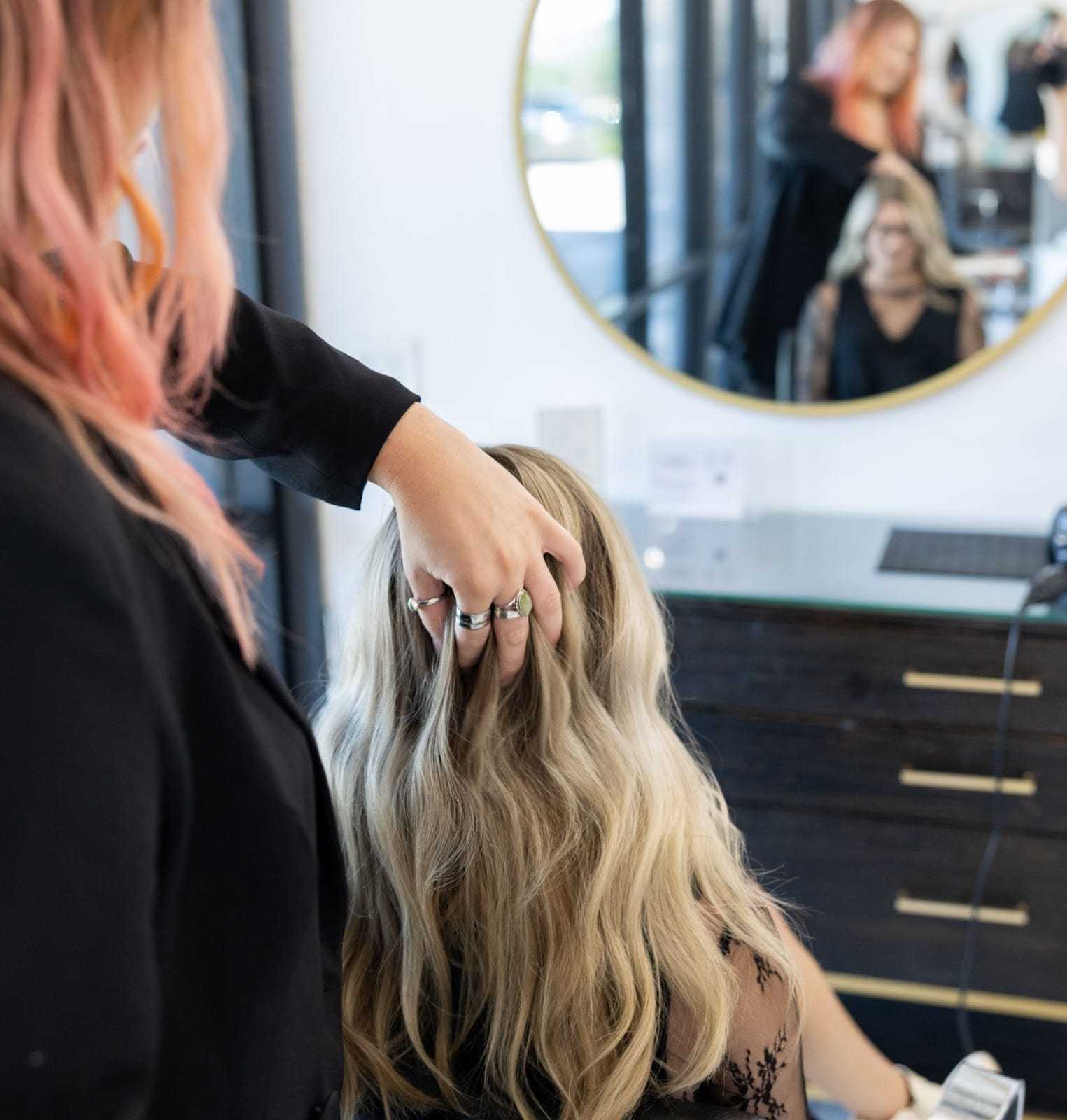 Hair stylist adjusting client's long, wavy blonde hair in a salon with a round mirror.